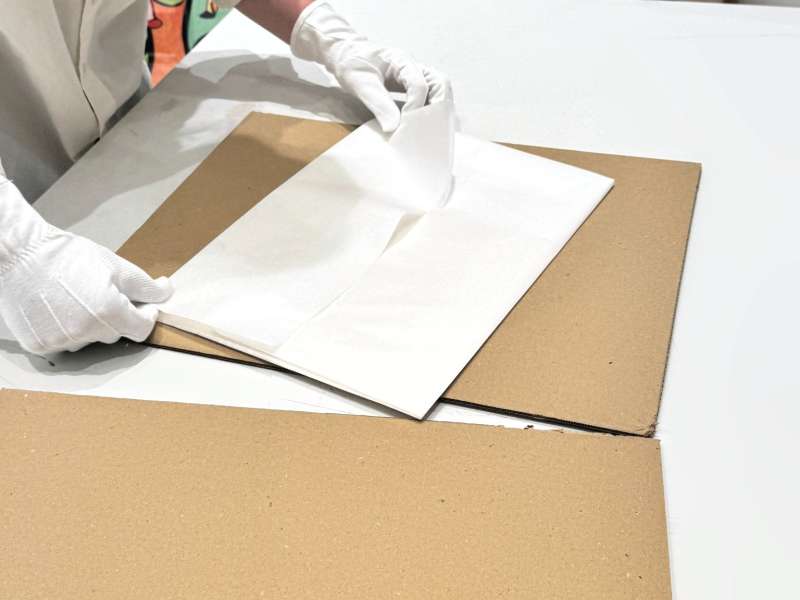 Préparation de l’emballage d’une estampe : feuilles de papier de soie posées sur un carton rigide, manipulées avec des gants de coton blancs